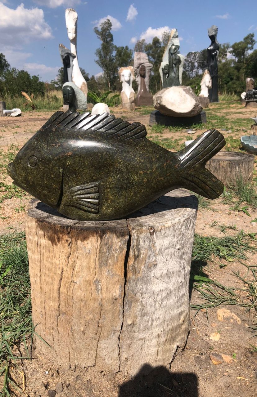 Polished stone fish sculpture with detailed fins and scales displayed on a wooden stump, surrounded by abstract outdoor sculptures under partly cloudy sky.