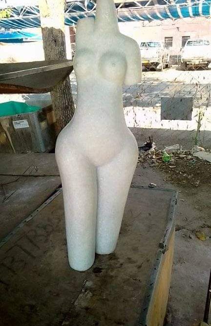 Light-colored humanoid mannequin standing on rugged surface, featuring a smooth texture and simplified human form without facial details, set against a blurred outdoor background.