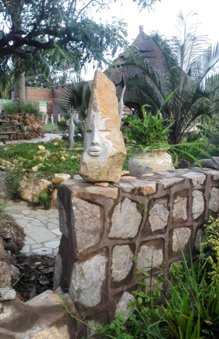 A stone sculpture resembling a face atop a rustic wall, surrounded by lush greenery in a tranquil garden.