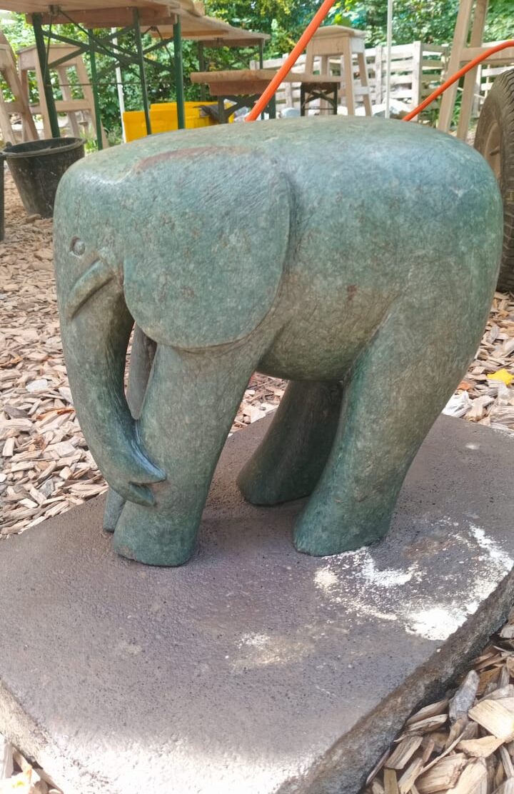Detailed sculpture of an elephant made from dark textured material, standing on a stone base with wooden tables in the background and sunlight filtering through greenery.