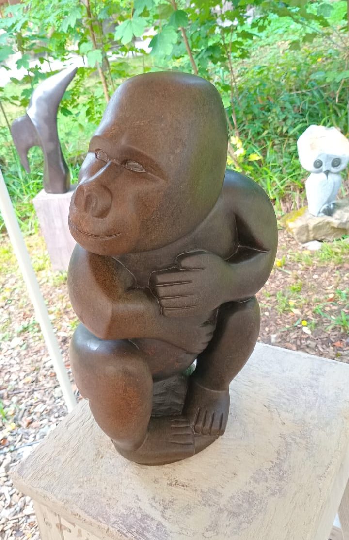 Detailed sculpture of a seated gorilla with arms wrapped around its knees, displayed on a rustic wooden table surrounded by greenery and additional sculptures in the background.