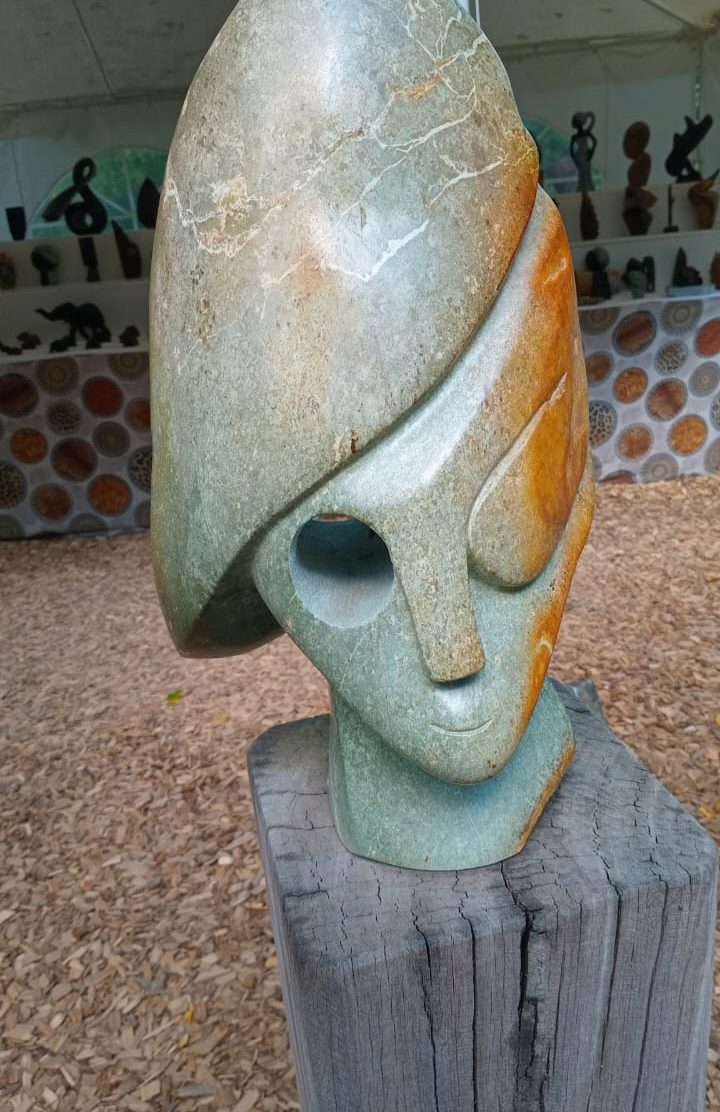 A contemporary stone sculpture representing a stylized face, designed with smooth gray and orange hues, situated on a weathered wooden pedestal amidst various art pieces in a tent-like environment, with shredded wood covering the ground.