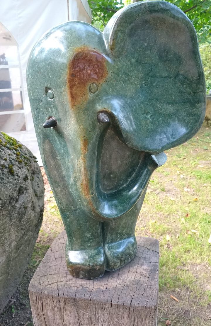 Green stone sculpture of a stylized elephant with a playful expression, standing on a wooden post amidst a blurred outdoor background of trees and a white tent.