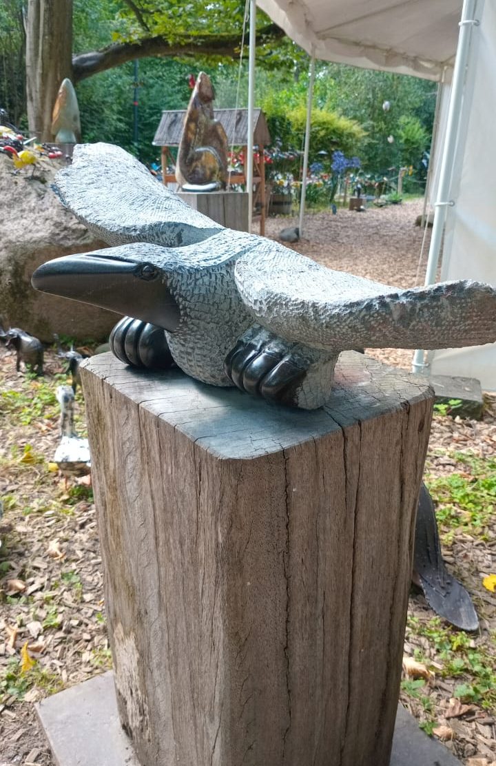 Detailed sculpture of a bird with outstretched wings perched on a rustic wooden pedestal, surrounded by lush greenery and artistic elements in a natural outdoor setting.