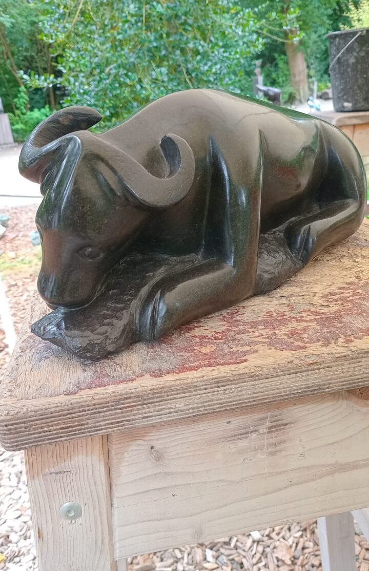 Bronze sculpture of a relaxed reclining cow with polished surface on a wooden table, surrounded by green foliage.