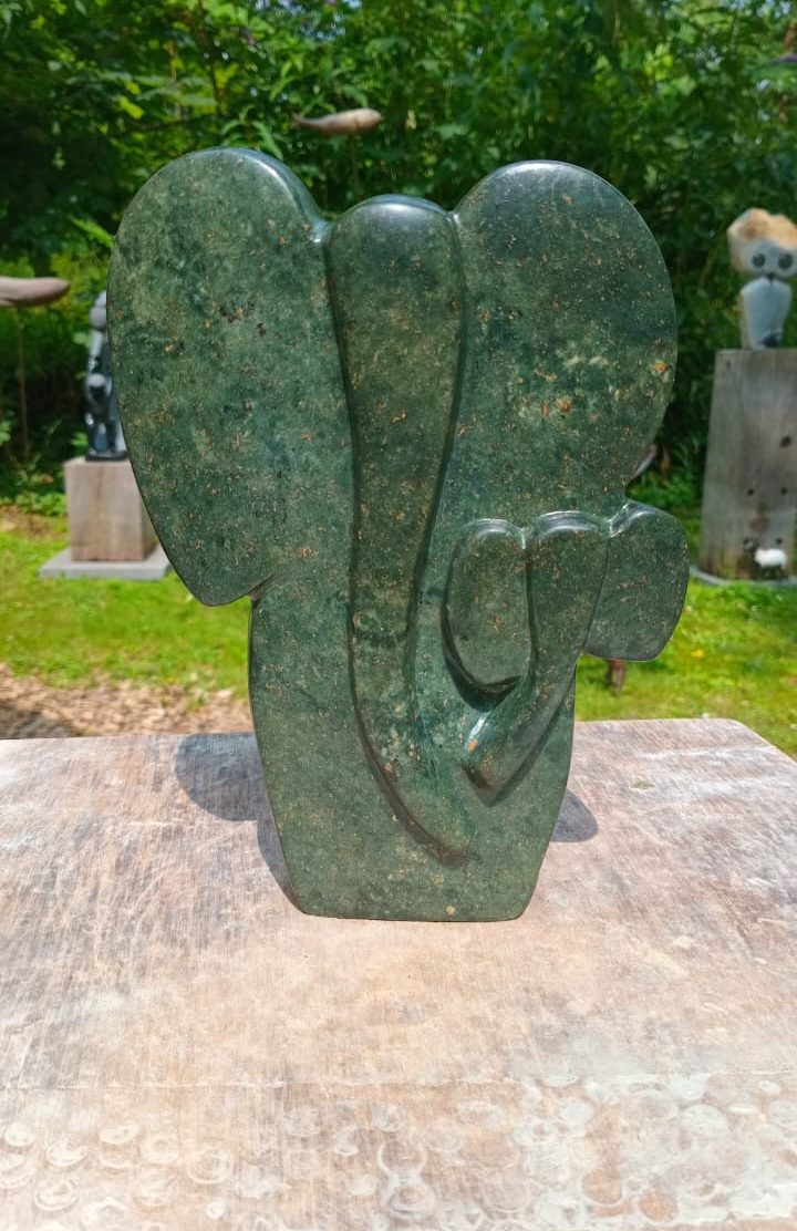A green stone sculpture of an abstract elephant with smooth curves, showcased on a wooden surface against a backdrop of lush greenery.