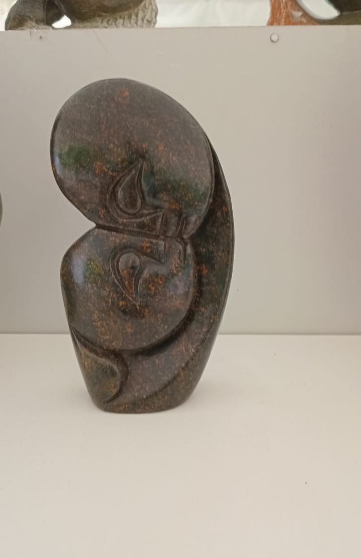 An abstract dark stone sculpture with a smooth, polished surface showcasing fluid, organic shapes and subtle textures, displayed on a plain background.