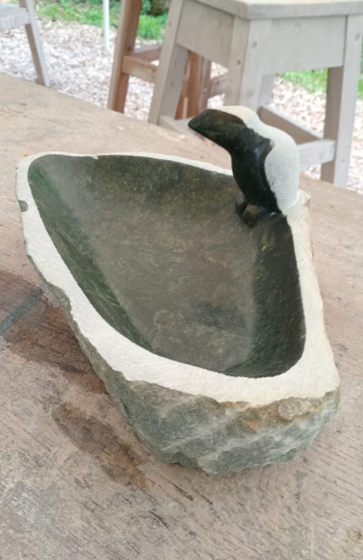 Sculpted stone bowl featuring a smooth interior and rough exterior, with a small bird figure on the rim, in a rustic workshop setting.