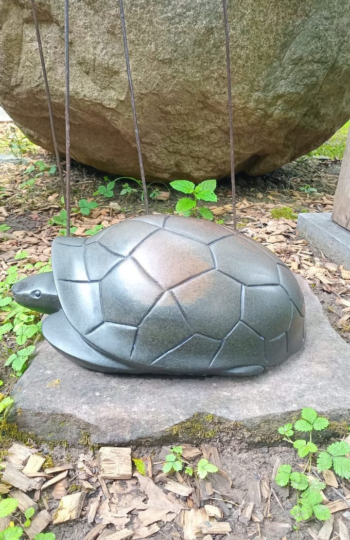 Polished stone turtle sculpture with detailed carvings resting on a flat stone surface surrounded by green plants and a large rock in a serene outdoor environment.