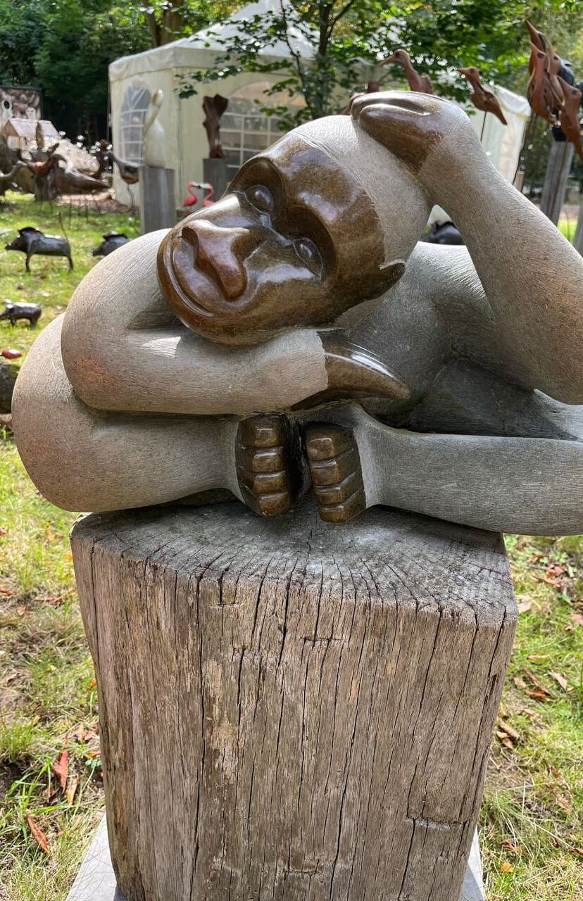 Stone sculpture of a gorilla resting its head on its hand, showcasing detailed facial features and a peaceful expression, set on a wooden pedestal in a serene garden.