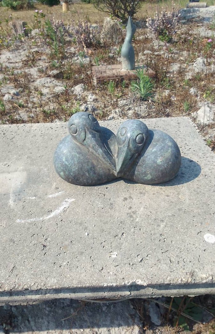 Close-up view of a unique sculpture featuring two stylized bird forms made of metallic material, set on a concrete slab with surrounding vegetation and distant sculptures.