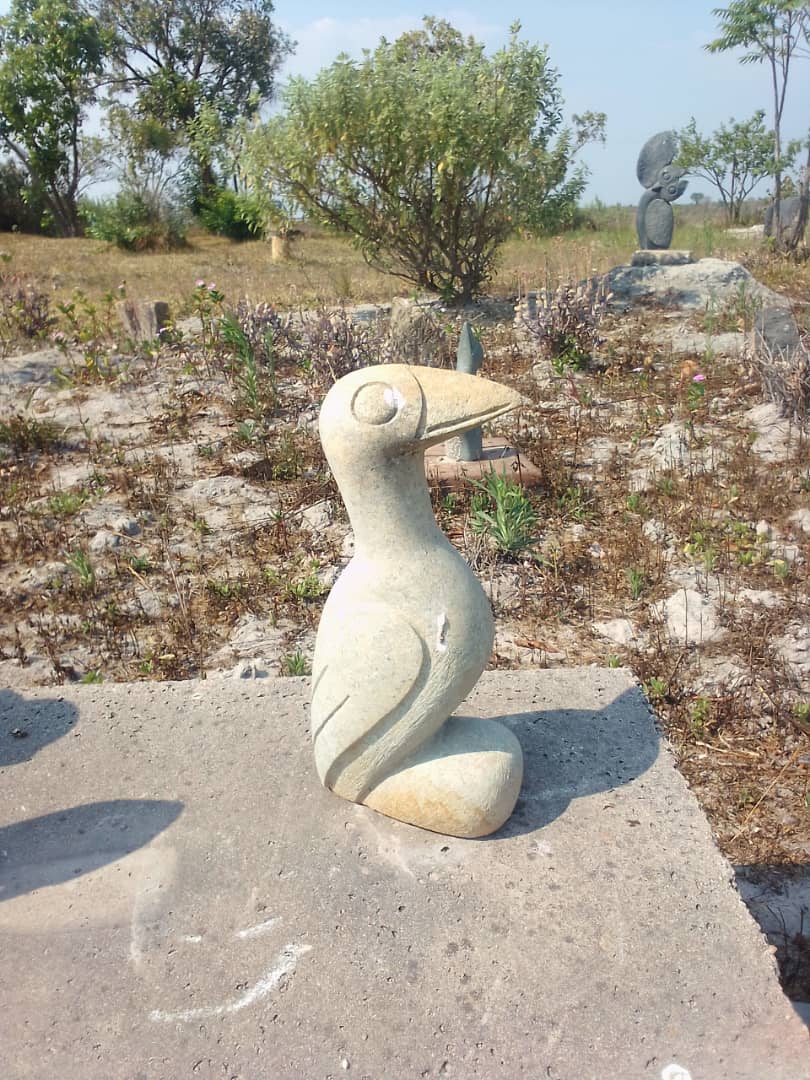 A carved stone sculpture of a sleek bird with a rounded head and pointed beak, set against a backdrop of sparse vegetation and a clear blue sky in an outdoor art installation.
