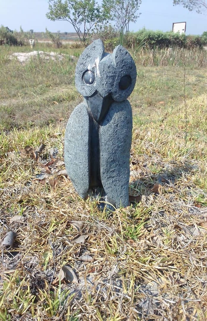 Textured owl sculpture with large eyes standing on grass surrounded by sparse vegetation and distant trees