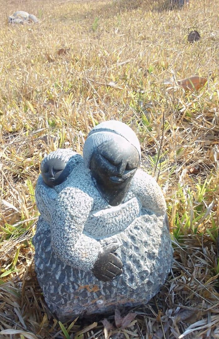 Textured stone sculpture depicting a woman carrying a child on her back, set against a backdrop of dry grass, emphasizing traditional design features.