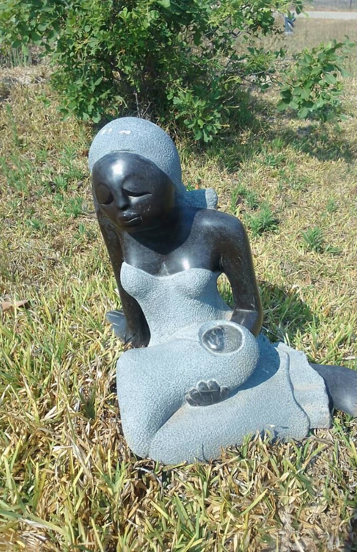 Bronze and stone sculpture depicting a serene seated woman holding a child in her lap amidst a tranquil grassy backdrop.