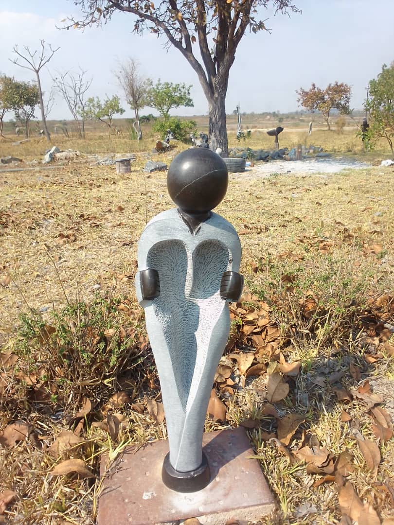 Unique abstract stone sculpture with heart-shaped torso and glossy head, surrounded by dry leaves and open grassy area under a clear blue sky.