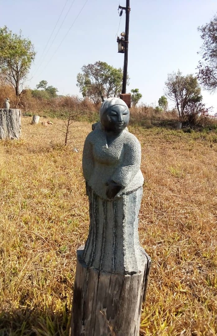 Artistic stone sculpture of a woman set in a rural grassy field, showcasing textured details and a traditional pose