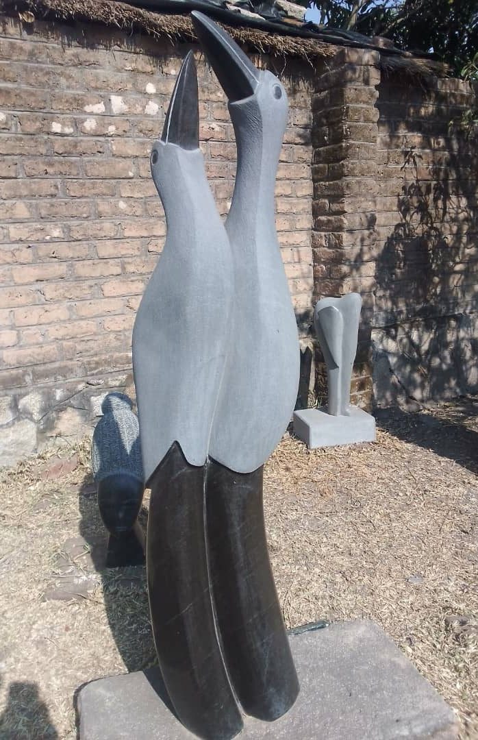 A striking sculpture of two stylized birds crafted from dark and light stone, standing side by side against a rustic brick wall with dry grass beneath, symbolizing a serene moment in nature.