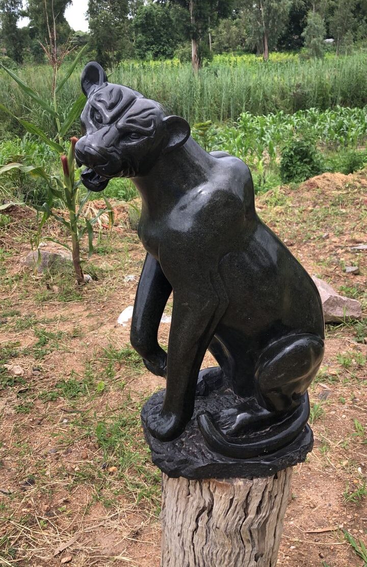 Detailed black stone sculpture of a large cat on a wooden stump, surrounded by lush greenery and earthy ground.