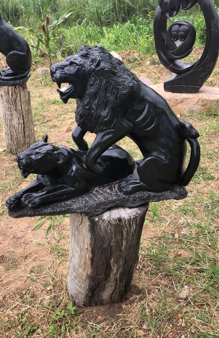 Detailed sculpture of a lion and a lioness carved from dark stone, featuring the lion in a powerful stance with an open mouth, and the lioness lying beneath; surrounded by greenery and abstract sculptures.