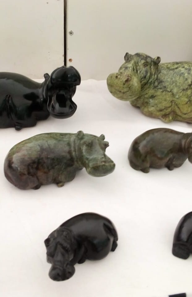 A variety of small hippo figurines in black, brown, and green, showcasing intricate designs on a white surface with a stone and wooden sculpture in the background.