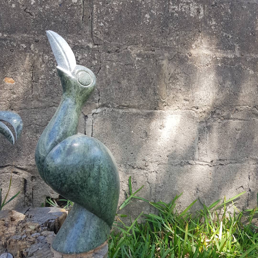 Sculpture of a bird made from green stone, arched neck and raised beak, set against a textured gray wall with sunlight highlighting its features, surrounded by fresh green grass.