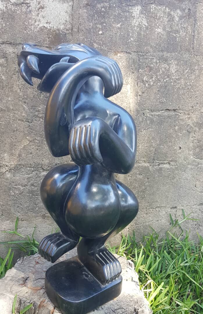 A black sculpture portraying a stylized figure with an elongated neck and exaggerated limbs, set against a textured gray background with grass in the foreground.