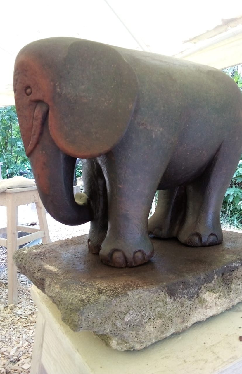 A textured sculpture of a cheerfully stylized elephant on a rectangular stone base, set in a shaded outdoor environment with greenery in the background.