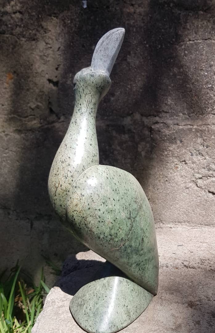 Smooth green stone sculpture of an African Fish Eagle - Hungwe - the Zimbabwean National Bird with a rounded body and elongated neck, set against a gray stone wall, featuring patches of sunlight and grass in the scene.
