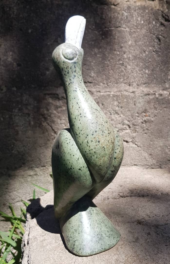 Stylized green stone sculpture of an African Fish Eagle - Hungwe - the Zimbabwean National bird with a white beak, placed on a stone surface, illuminated by natural light against a textured wall and grass.