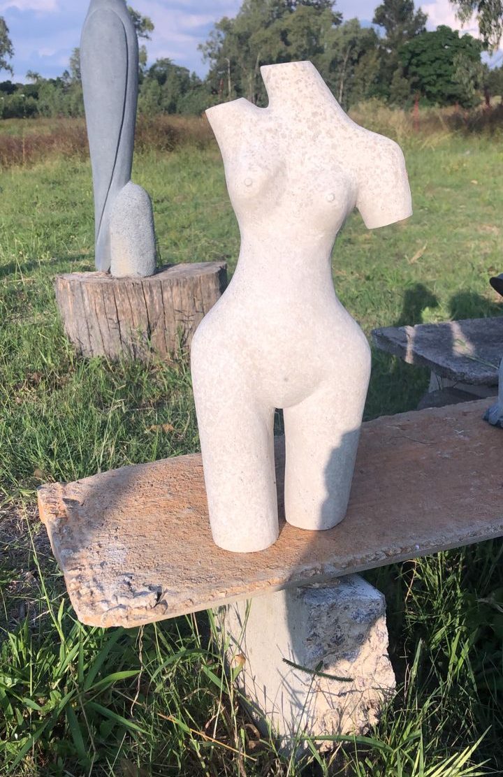 A light-colored stone sculptural representation of a stylized torso displayed on a rustic wooden bench outdoors, surrounded by greenery and natural elements under a clear blue sky.