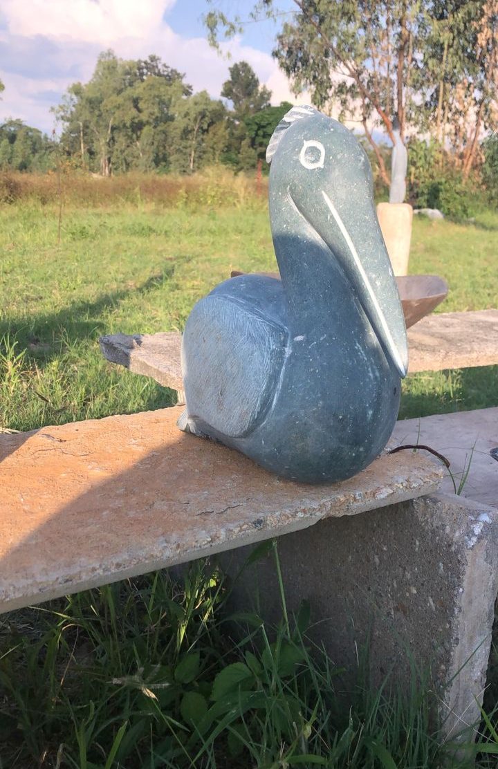 A beautifully carved stone sculpture resembling a pelican, with a bluish-grey color and white accents, set against a tranquil outdoor background of lush greenery and a blue sky.