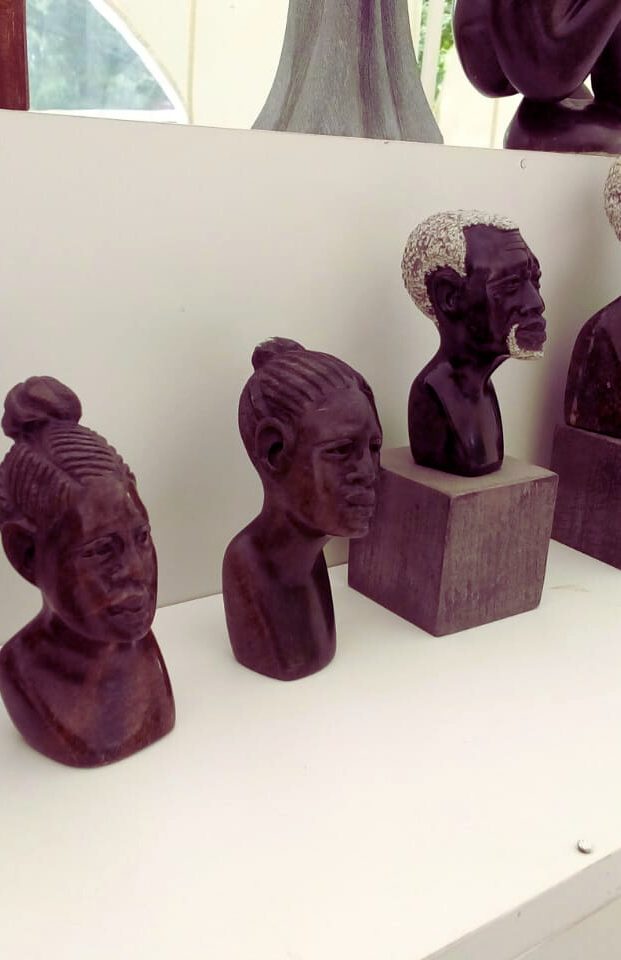 Collection of handcrafted wooden sculptures depicting human busts with various hairstyles and facial expressions, displayed on a white shelf under soft natural light.