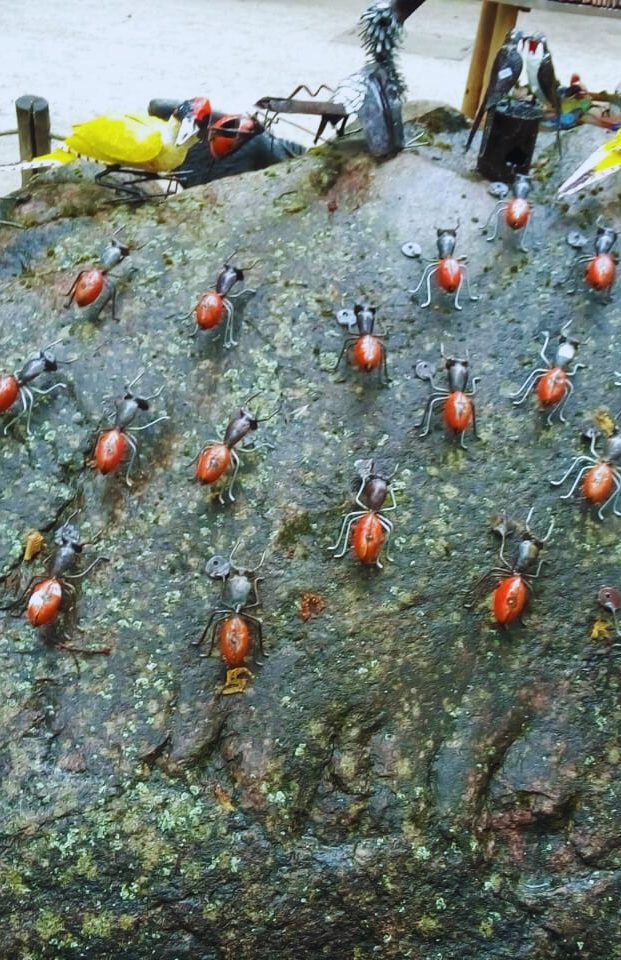 Textured rock decorated with clusters of black and reddish ant sculptures in an outdoor environment, with blurred artistic birds in the background.