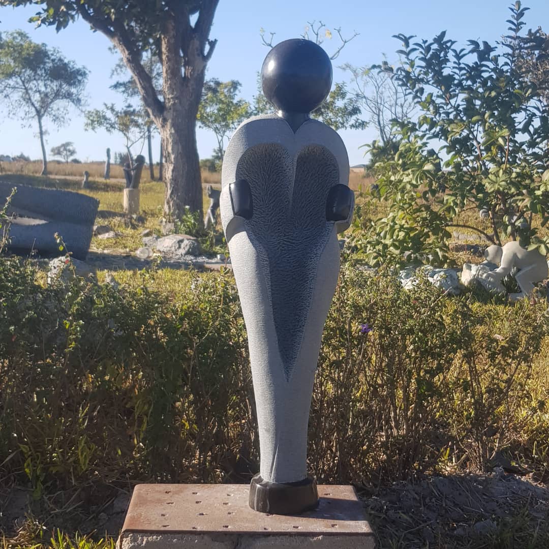 A striking gray sculpture with a heart shape at its center and a black spherical head, surrounded by grass and shrubs under a clear blue sky.