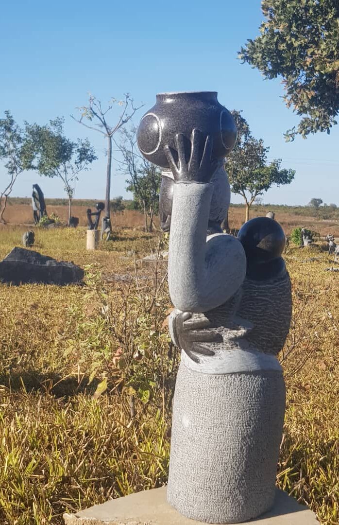 Carved stone sculpture of a rounded figure with one arm raised, holding a pot above its head, set against a serene outdoor landscape with grassy fields and scattered trees.