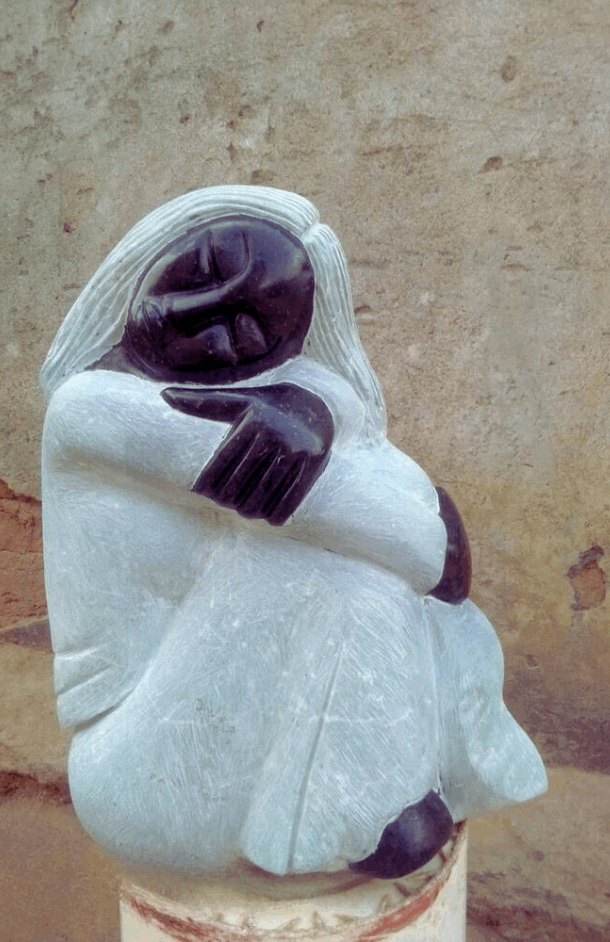 Close-up of a beautifully carved sculpture depicting a seated figure with a serene expression, made of dark stone and translucent light stone, hugging its knees against a textured wall.