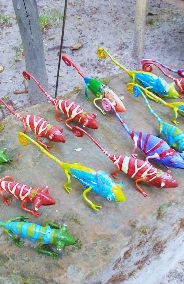 Vibrant handcrafted lizard figurines in red, green, blue, and yellow displayed on a natural stone surface, surrounded by greenery.